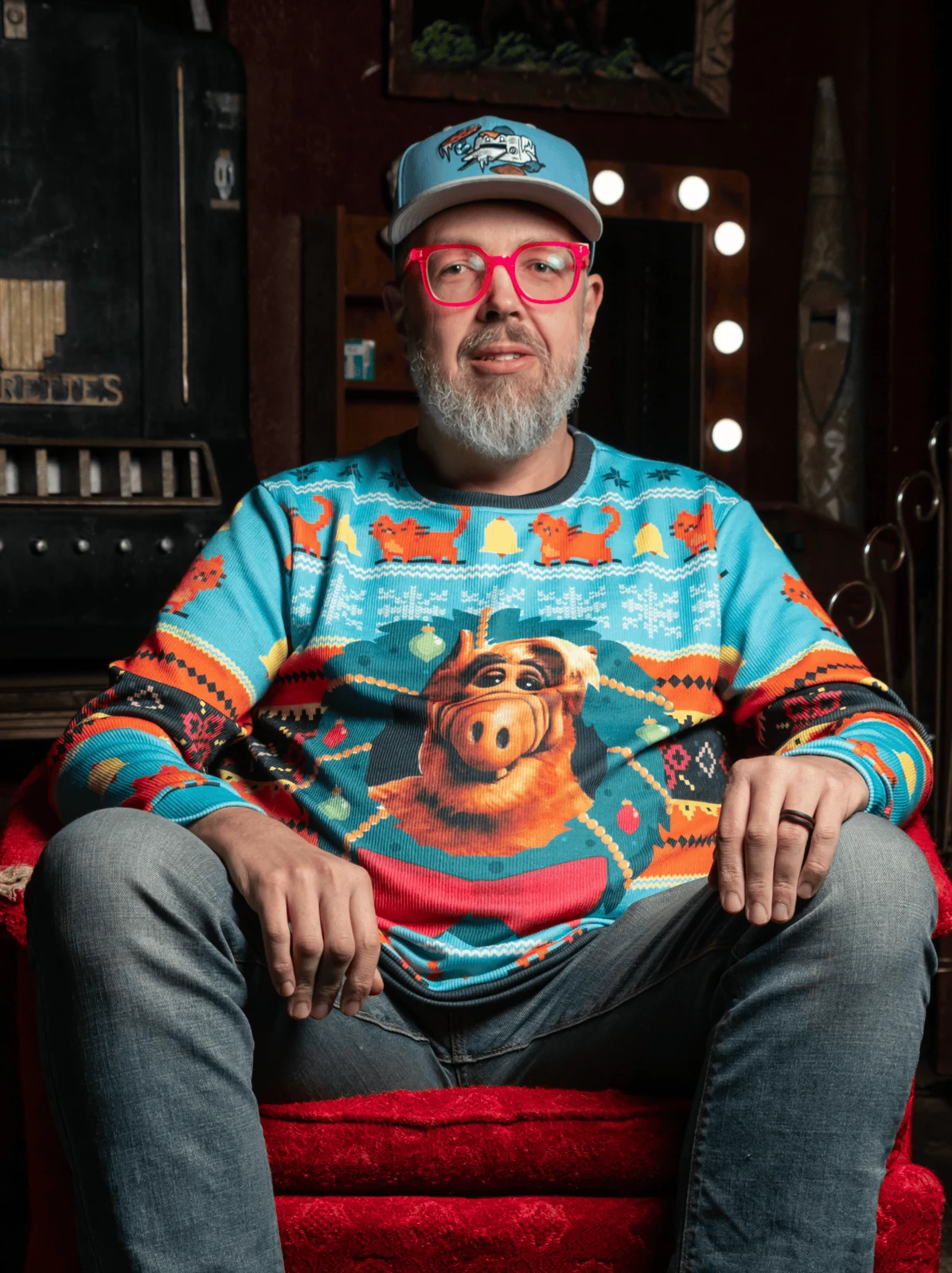 Michael Charron seated in a red chair wearing colorful glasses, a cap, and a Muppets sweater
